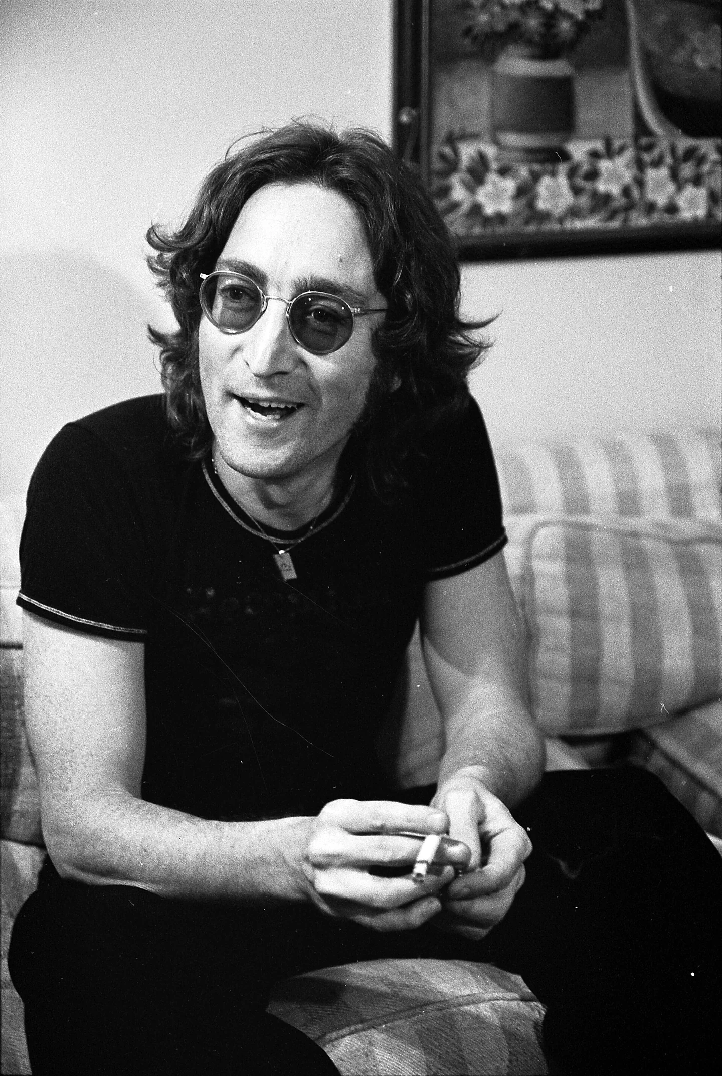 John Lennon in round glasses, sitting on a sofa and leaning forward as he talks (black-and-white photo)